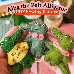 May include: Three handmade felt alligators in shades of green are shown. One has a pink bow tie, another a yellow banner reading "SEE YOU LATER", and the third a pink scarf. The text "Alba the Felt Alligator PDF Sewing Pattern" is at the top of the image.