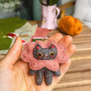 May include: A handmade felt cat brooch with a pink flower-shaped collar. The gray cat has yellow eyes and red accents. The brooch is held in a hand, with a wooden table and flowers in the background.