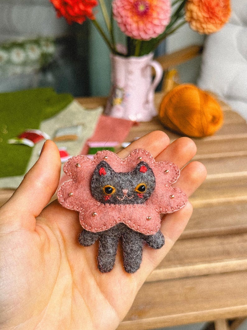 May include: A handmade felt cat brooch with a pink flower-shaped collar. The gray cat has yellow eyes and red accents. The brooch is held in a hand, with a wooden table and flowers in the background.
