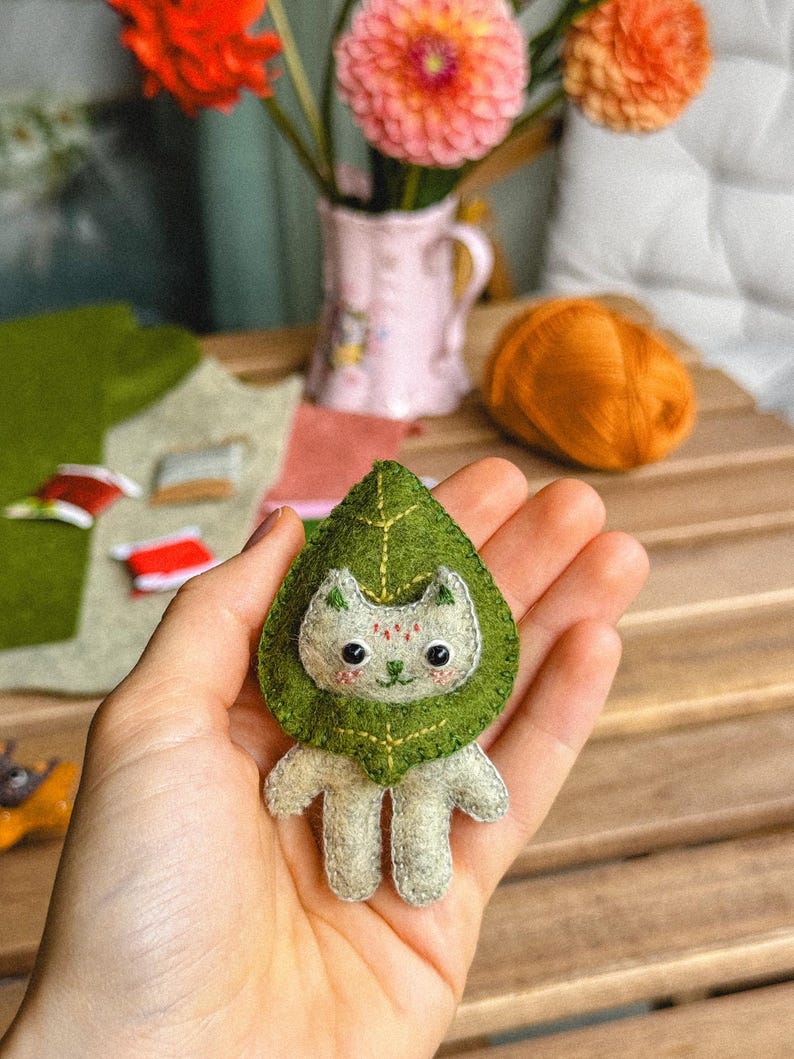 May include: A handmade felt cat figurine with a green leaf-shaped collar. The cat is light gray with embroidered details, including a smiling face and small red accents. The background includes flowers in a vase and a ball of orange yarn.