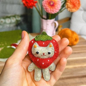 May include: A handmade felt cat-shaped pin, designed to resemble a strawberry. The cat has a white face, gray body, and is surrounded by a red strawberry with white dots and a green leaf top. The pin is held in a hand.