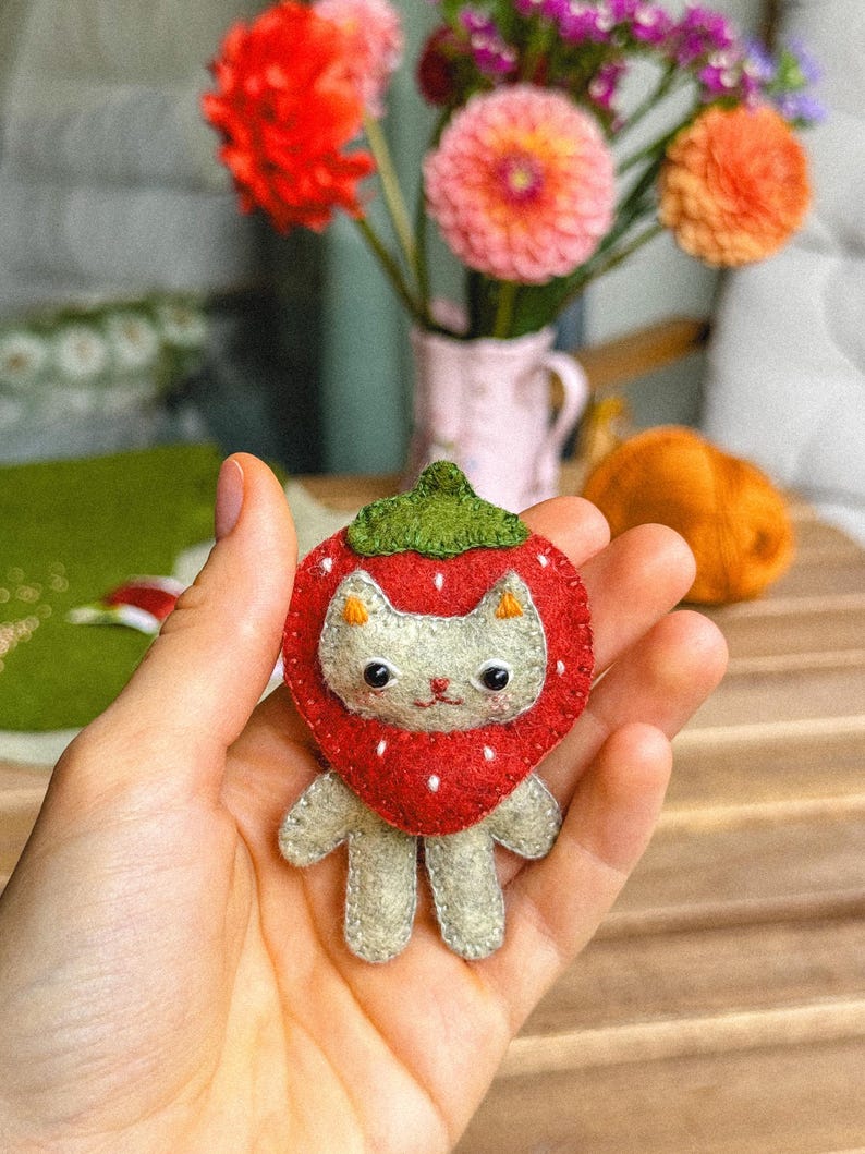 May include: A handmade felt cat-shaped pin, designed to resemble a strawberry. The cat has a white face, gray body, and is surrounded by a red strawberry with white dots and a green leaf top. The pin is held in a hand.