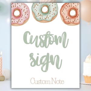 May include: A white sign featuring three watercolor donuts in pink, green, and peach with sprinkles. The words "Custom sign" are written in a soft green script, with "Custom Note" in a smaller, brown font below. The sign is likely for a party or event.