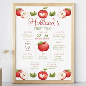 May include: A framed print with a wooden frame, featuring a watercolor design with apples and leaves. The print includes the text "Holland's FIRST YEAR", along with details about the child's first year, including weight, height, favorite toys, and foods.