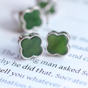 May include: Close-up of two silver-toned clover-shaped stud earrings with a dark green center. The earrings are on a white surface with printed text. The earrings are a jewelry item.