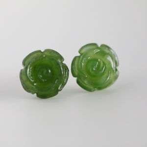 May include: Two green jade rose-shaped stud earrings. The earrings are carved with detailed petals and a swirling center. The jade has a translucent quality, and the earrings are set against a white background.