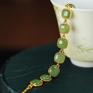 Könnte beinhalten: Ein goldfarbenes Armband mit quadratischen, hellgrünen Jadesteinen. Die Steine sind in goldfarbenen Fassungen gefasst und durch zarte goldene Glieder verbunden. Das Armband wird vor einem dunkelgrünen Hintergrund gezeigt.