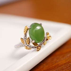 Op de afbeelding: Een zilveren ring met een grote, ovale, groene edelsteen. De ring heeft een goudkleurig bladerontwerp rond de steen. De ring ligt op een wit oppervlak met een vage achtergrond.