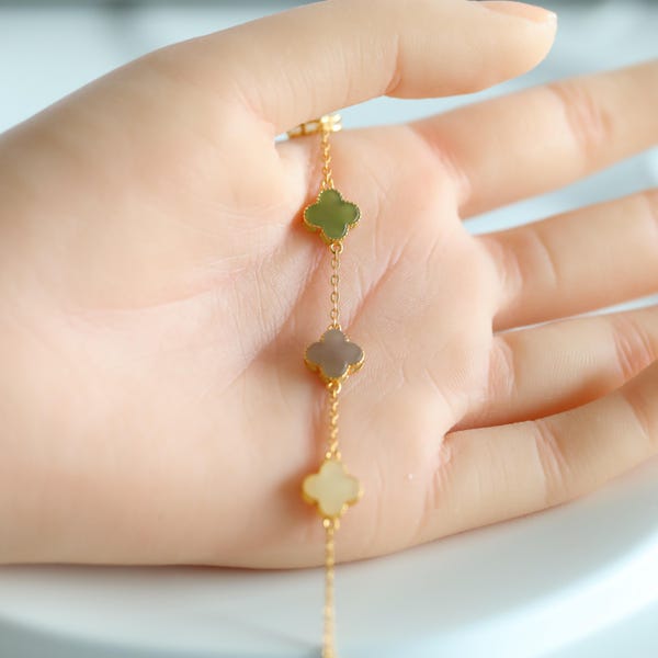 May include: A gold-colored chain bracelet featuring three clover-shaped charms in shades of green, gray, and cream. The bracelet is delicate and elegant, with each charm connected by a fine chain. The bracelet is a piece of jewelry.