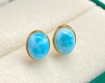 May include: A pair of gold-rimmed stud earrings featuring oval, light blue gemstones. The earrings are set against a white background inside a green jewelry box, showcasing the vibrant blue of the stones.