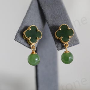 May include: A pair of gold-toned earrings featuring a four-leaf clover design with a green jade stone center. Each earring has a dangling, round green jade bead. The earrings are displayed on a gray surface.