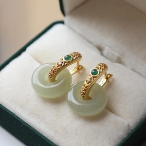 Op de afbeelding: Een paar goudkleurige oorringen met een lichtgroene, cirkelvormige jade hanger. De oorbellen hebben een decoratieve gouden sluiting met een kleine groene steen en verschillende kleine heldere stenen. Gepresenteerd in een groene sieradendoos.