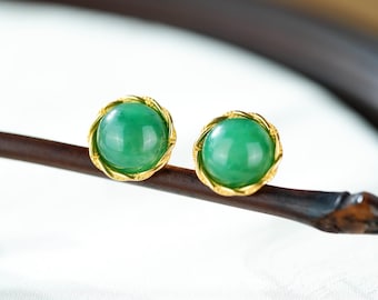 Könnte beinhalten: Ein Paar runde, grüne Jade-Ohrringe mit goldfarbenen, gedrehten, seilartigen Fassungen. Die Ohrringe sind vor einem verschwommenen Hintergrund platziert, der die leuchtende Farbe und das Design des Schmucks hervorhebt.
