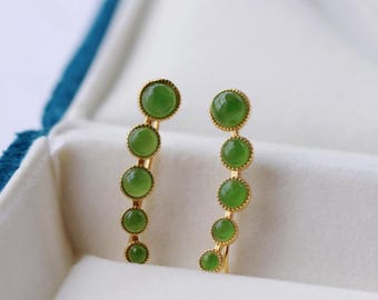 Könnte beinhalten: Ein Paar goldene Ohrringe mit jeweils fünf grünen Jade-Steinen. Die Ohrringe sind gebogen und die Steine sind in einer goldenen Fassung gefasst.