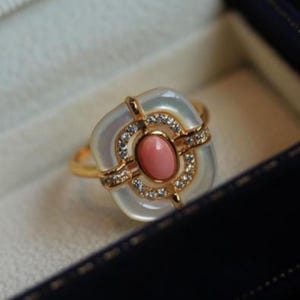 May include: A gold ring featuring a central oval pink gemstone, surrounded by a square, iridescent white stone. The ring is accented with small, sparkling clear stones and set in a gold band. The ring is displayed in a dark blue jewelry box.
