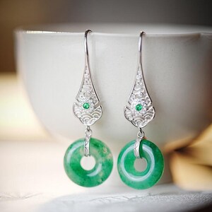 Può includere: Un paio di orecchini pendenti in argento con un design floreale a filigrana. Ogni orecchino presenta una piccola gemma verde e un pendente circolare di giada verde brillante. Sfondo sfocato.
