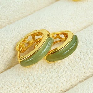 May include: A pair of gold-coloured hoop earrings with a green, oval-shaped inlay. The earrings have a decorative openwork design on the top portion. The earrings are displayed on a white, textured surface.