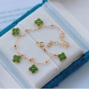 May include: A gold-tone bracelet with four-leaf clover pendants, each with a green gemstone centre. The bracelet has a fine chain with small gold beads and a clasp. It is presented in a blue velvet jewellery box.