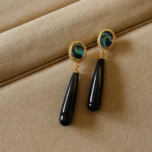 May include: A pair of gold-coloured earrings with a black, teardrop-shaped dangle. The earrings feature an oval-shaped top with a green and blue iridescent stone. The earrings are displayed on a tan surface.