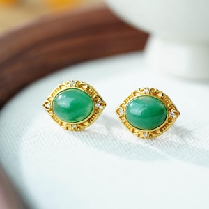 May include: A pair of gold-colored stud earrings with oval, bright green gemstones. The earrings have a decorative filigree border with small, clear accent stones. The earrings are displayed on a white surface.