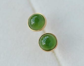 Könnte beinhalten: Ein Paar Ohrstecker mit runden, leuchtend grünen Edelsteinen, die in goldfarbenen Fassungen gefasst sind. Die Ohrringe haben eine polierte Oberfläche und reflektieren das Licht. Der Schmuck wird auf einer hellen Oberfläche präsentiert.