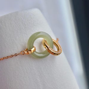 May include: A delicate gold-toned bracelet featuring a light green jade ring interlocked with a smaller, diamond-studded gold ring. The jade ring is smooth and circular, while the other ring is a curved shape. The bracelet chain is fine and elegant.