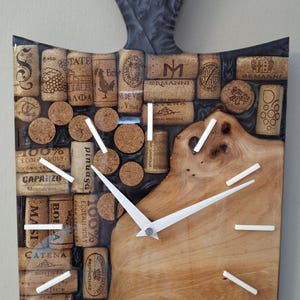 May include: A unique wall clock made from wine corks and wood. The clock face is a light brown wood with white hands and markers. The handle is a dark gray, and the clock is filled with various wine corks.