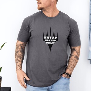 May include: A dark gray t-shirt with a black and white graphic of a five-pointed star with the text "UNTAP UPKEEP DRAW" underneath.