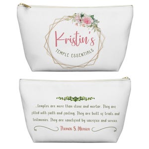 Op de afbeelding: Witte make-uptas met een gouden rits. De tas heeft de tekst "Kristin's TEMPLE ESSENTIALS" in een gouden geometrische lijst met roze bloemen. De onderkant van de tas heeft een citaat van Thomas S. Monson.