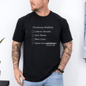 May include: Black t-shirt with a white Christmas wishlist graphic. The text includes "Collector Booster," "Deck Sleeves," "Black Lotus," and "Spend time with family playing magic."