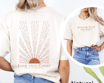 Puede incluir: Una camiseta blanca con un diseño de sol marrón y el texto "Look Unto Christ, Doctrine and Covenants 88:63" impreso en la espalda. La camiseta se muestra en una modelo que lleva pantalones vaqueros azules.