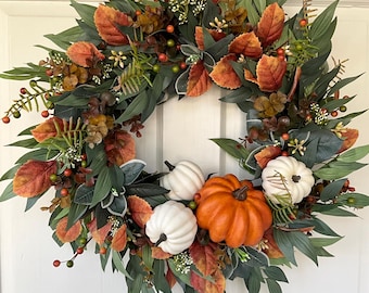 May include: A fall-themed wreath with a mix of green, orange, and red leaves, berries, and small white and orange pumpkins. The wreath is circular and designed for a door or wall decoration. The wreath is set against a white door.