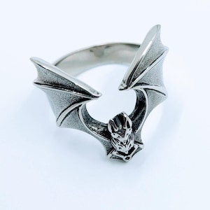 Peut inclure: Une bague en argent avec un motif de chauve-souris. Les ailes de la chauve-souris sont déployées de chaque côté de la bague, et le corps de la chauve-souris est au centre de la bague.