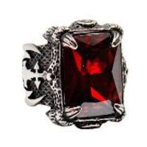 May include: A silver ring featuring a large, red, rectangular gemstone set in a claw setting. The ring has an intricate design with a cross on the side.