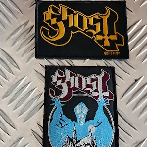 Könnte beinhalten: Zwei schwarze, gestickte Aufnäher mit der Band Ghost. Ein Aufnäher hat goldfarbene Schrift und ein Kreuzsymbol. Der andere Aufnäher hat rote und weiße Schrift, eine blaue Figur und eine Kirchenillustration.