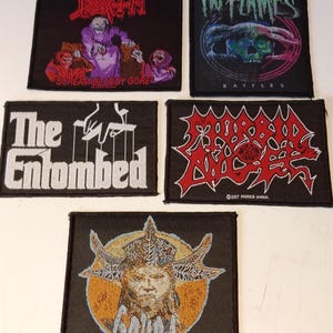 May include: Five black fabric patches with band logos. The patches feature the logos of Death, In Flames, The Entombed, Morbid Angel, and Gojira. The Gojira patch has a detailed illustration of a figure with horns. The patches are rectangular and have embroidered designs.