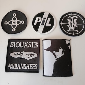 Puede incluir: Colección de parches bordados en blanco y negro. Incluye un parche redondo con "PiL" en blanco, un parche rectangular con el texto "Siouxsie and the Banshees" y una silueta de una persona con sombrero.