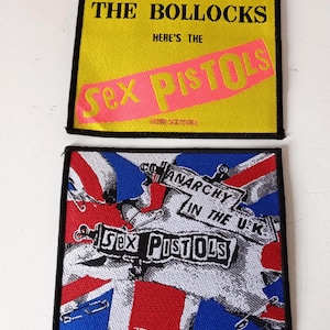 May include: Two square patches with black borders. The top patch is yellow with black text that reads "NEVER MIND THE BOLLOCKS" and "Sex Pistols" in pink. The bottom patch features a Union Jack design with "Sex Pistols" and "Anarchy in the UK" text.