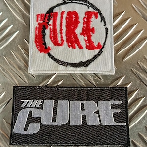 Puede incluir: Dos parches bordados con los logotipos de la banda "THE CURE". Uno es cuadrado con texto rojo dentro de un círculo negro sobre un fondo gris claro. El otro es rectangular con texto gris sobre un fondo negro.