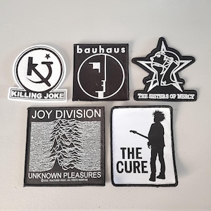 May include: A collection of nine black and white embroidered patches featuring band logos. The patches include Killing Joke, Bauhaus, The Sisters of Mercy, Joy Division, and The Cure.