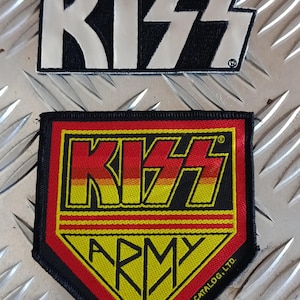 Peut inclure: Deux écussons brodés représentant le groupe KISS. Le patch supérieur est noir avec des lettres blanches. Le patch inférieur est rouge, jaune et noir, avec les mots "KISS ARMY" et le logo du groupe.