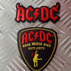 Könnte beinhalten: Zwei AC/DC-Patches. Der obere Patch zeigt das Logo der Band in Rot mit schwarzem Rand. Der untere Patch ist schildförmig, mit dem AC/DC-Logo, dem Text "ROCK NEVER DIES EST.1973" und einer gelben Silhouette einer Figur.