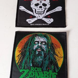 Pode incluir: Dois patches pretos com designs de Rob Zombie. Um apresenta uma caveira e ossos cruzados com o texto "Rob Zombie" em vermelho. O outro mostra um retrato de Rob Zombie com cabelo e barba verdes.