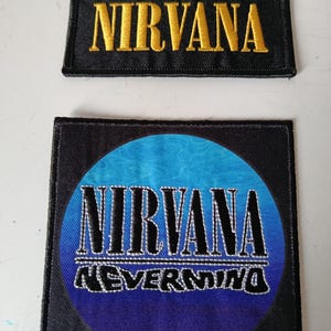 Peut inclure: Deux écussons en tissu noir. Le patch supérieur est rectangulaire avec le mot "NIRVANA" brodé en or. Le patch inférieur est carré avec un motif circulaire bleu et violet et les mots "NIRVANA NEVERMIND".