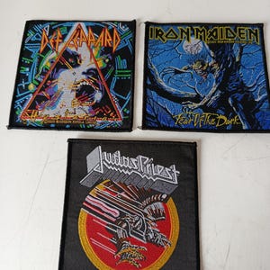 Puede incluir: Tres parches bordados con logotipos de bandas de rock. Un parche muestra "Def Leppard" con un diseño geométrico. Otro muestra "Iron Maiden" con una escena oscura e ilustrada. El tercer parche presenta "Judas Priest" con un gráfico rojo y amarillo.