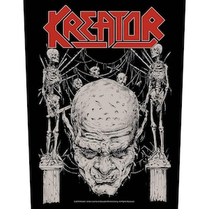 以下が含まれることがあります： 赤色の「KREATOR」の文字が描かれた黒色のグラフィックパッチ。イラストは、大きなグロテスクな顔と、骸骨とクモの巣に囲まれた、目立つ眉と歯が特徴です。パッチは黒色の背景で、詳細なイラストが描かれています。