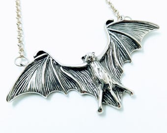 May include: A silver bat pendant necklace with a detailed design. The bat is in flight with its wings spread wide. The necklace is made of a silver chain.