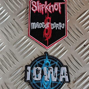 May include: Two embroidered patches featuring the band Slipknot. The top patch is a black hexagon with the band's name in red and the text "MAGGOT CORPS". The bottom patch is circular, with "IOWA" in white and a blue design.