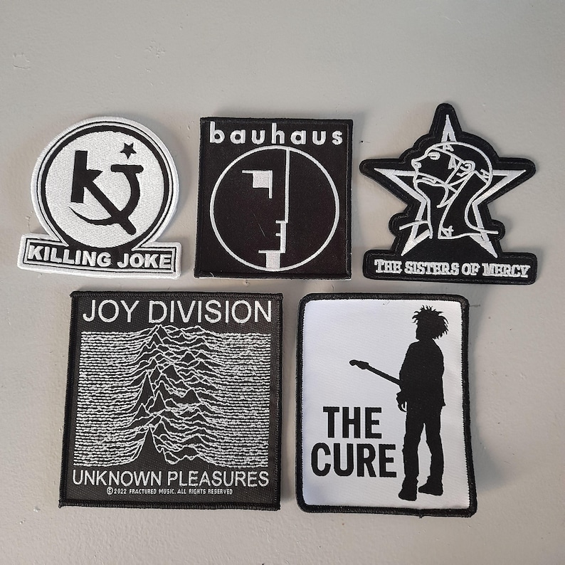 May include: Five black and white embroidered patches featuring the band names Bauhaus, Killing Joke, The Sisters of Mercy, Joy Division, and The Cure. The patches are all rectangular except for The Sisters of Mercy patch, which is a star shape.
