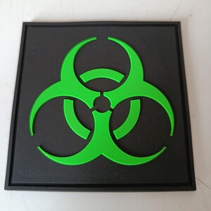 May include: A square black patch featuring a raised, bright green biohazard symbol centred on the surface. The design consists of three interlocking curved segments surrounding a central circle, all rendered in a bold, high-contrast colour scheme.